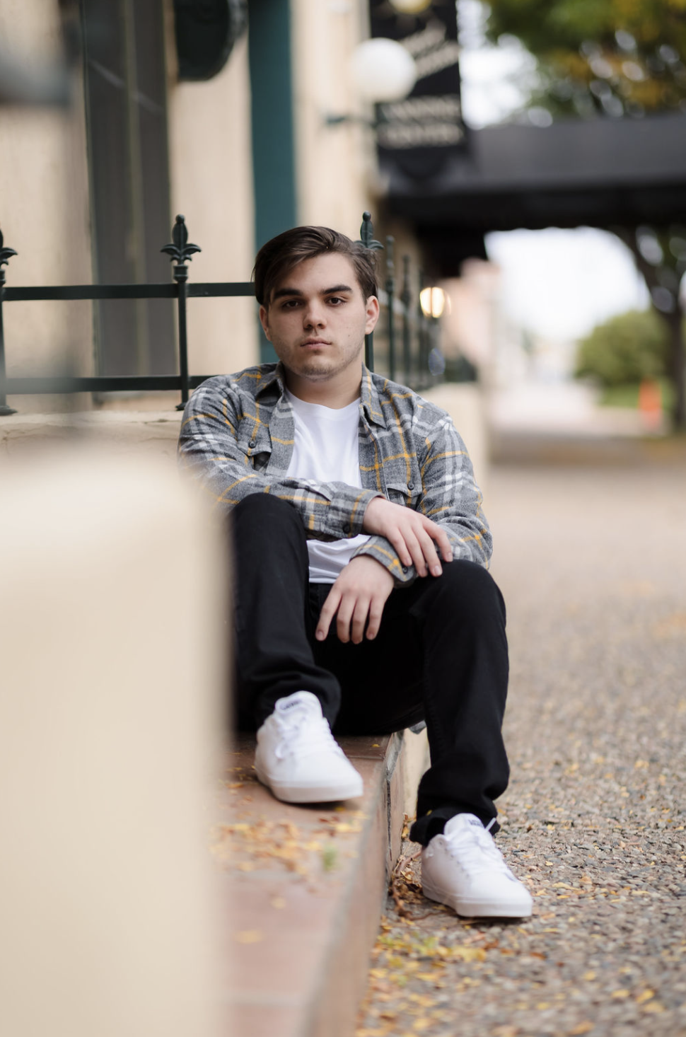 A young man with dark hair wearing a plaid shirt, white t-shirt, black pants, and white sneakers sitting on a sidewalk curb with a serious expression.