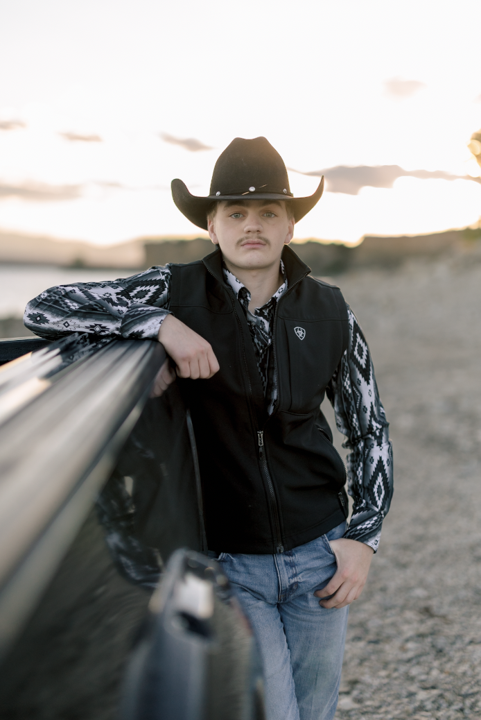 A young man in cowboy hat and patterned shirt leaning on a black car outdoors at sunset.