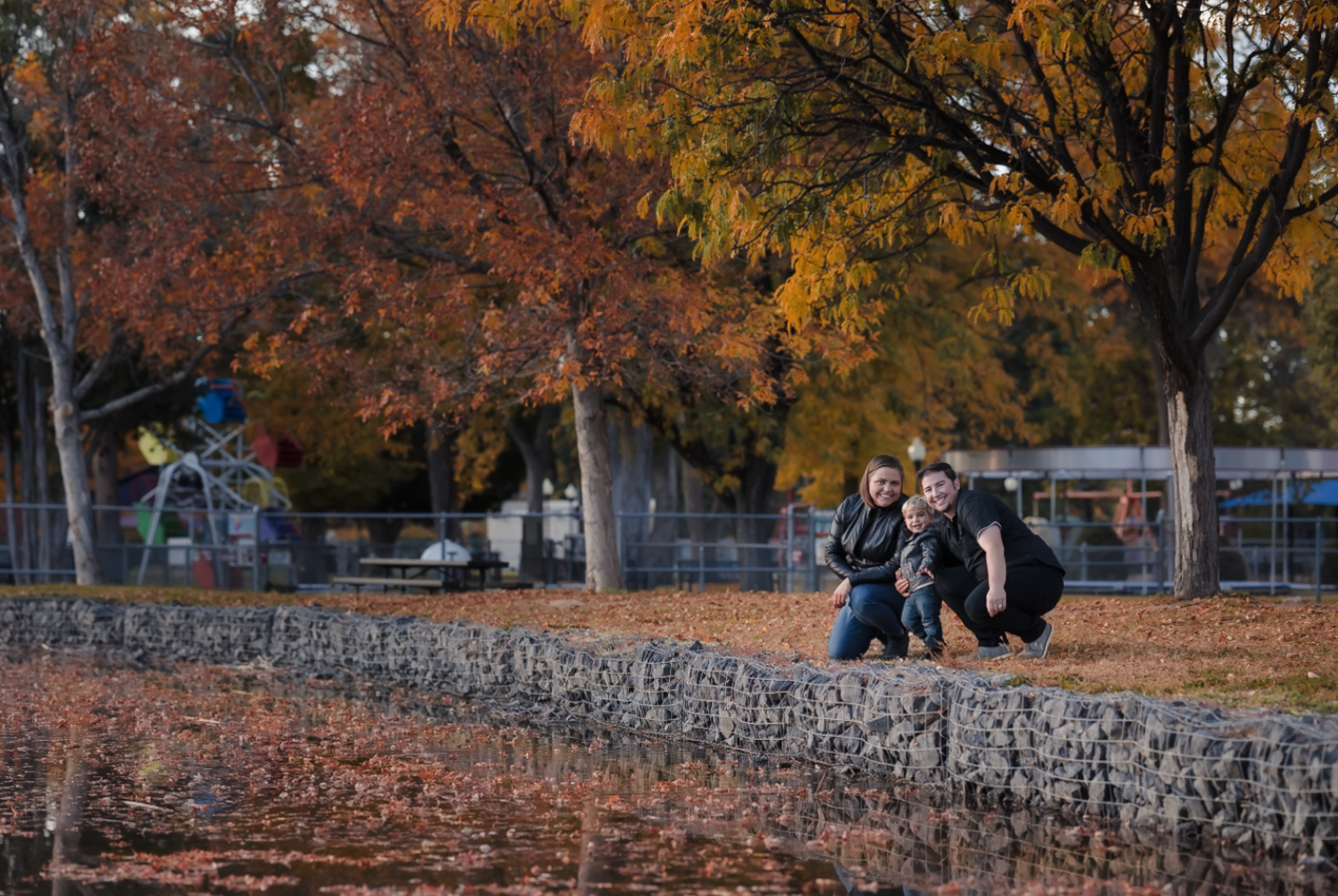 A young woman with dark brown wavy hair and blue eyes smiling outdoors in a park during daytime, wearing a white sweater and layered gold necklaces.