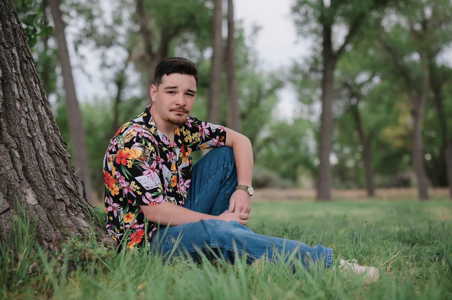 A young man sitting on the grass beside a tree in a park, wearing a colorful Hawaiian shirt and jeans, with a neutral expression, surrounded by trees and greenery.
