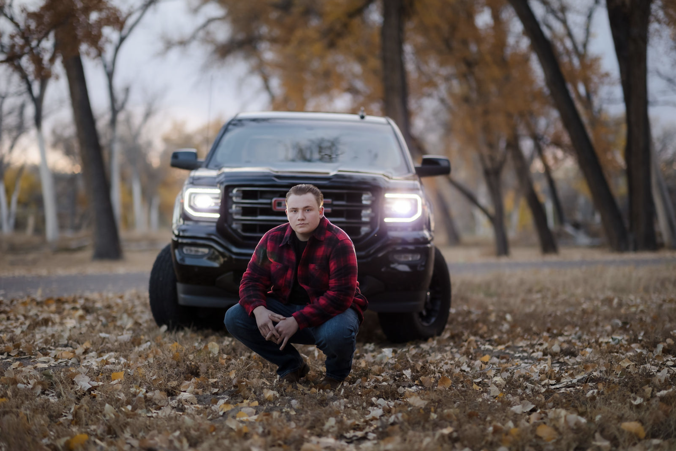 A young man crouches in a fall park with leaves on the ground, in front of a black truck with its headlights on, surrounded by trees with autumn-colored leaves.