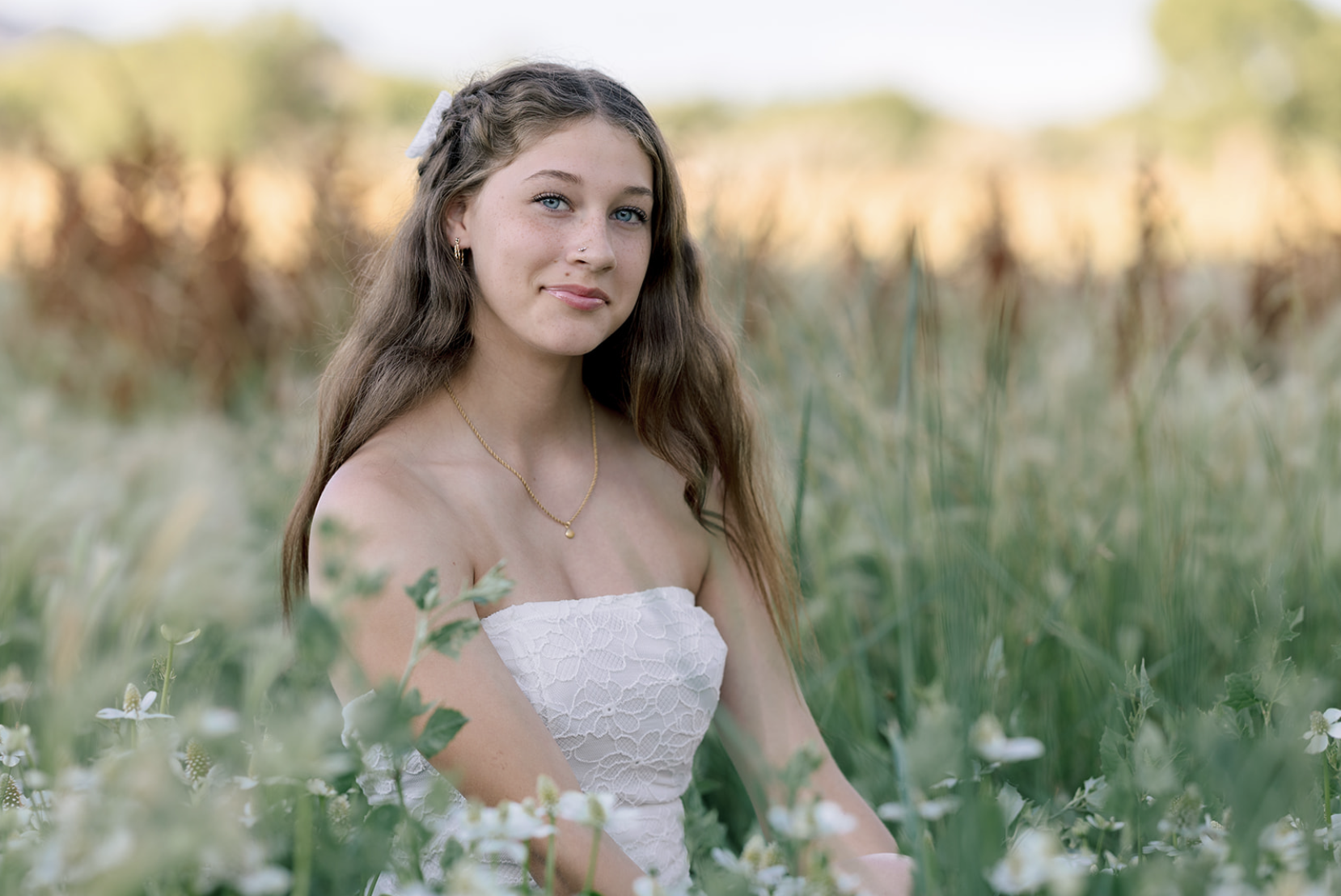 A young woman with long, wavy brown hair and blue eyes sitting in a green field with white flowers, wearing a white strapless dress and a gold necklace.