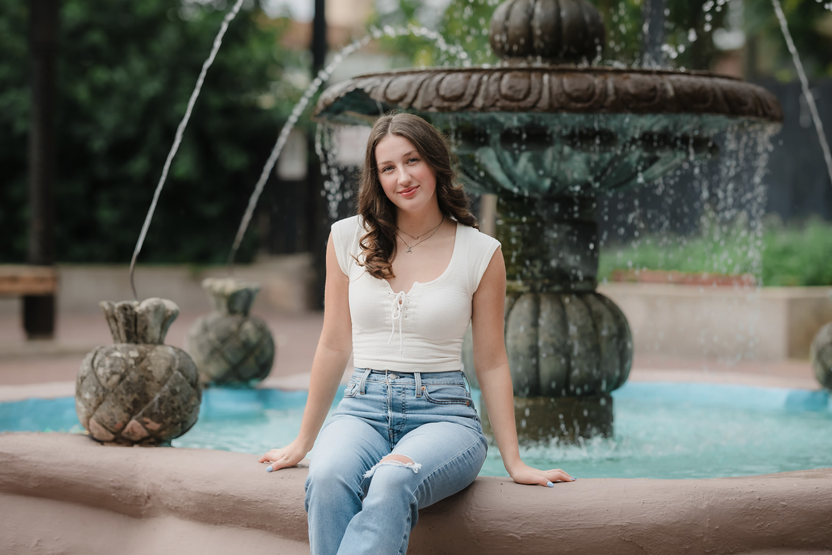 A young woman sitting on the edge of a fountain in a park.