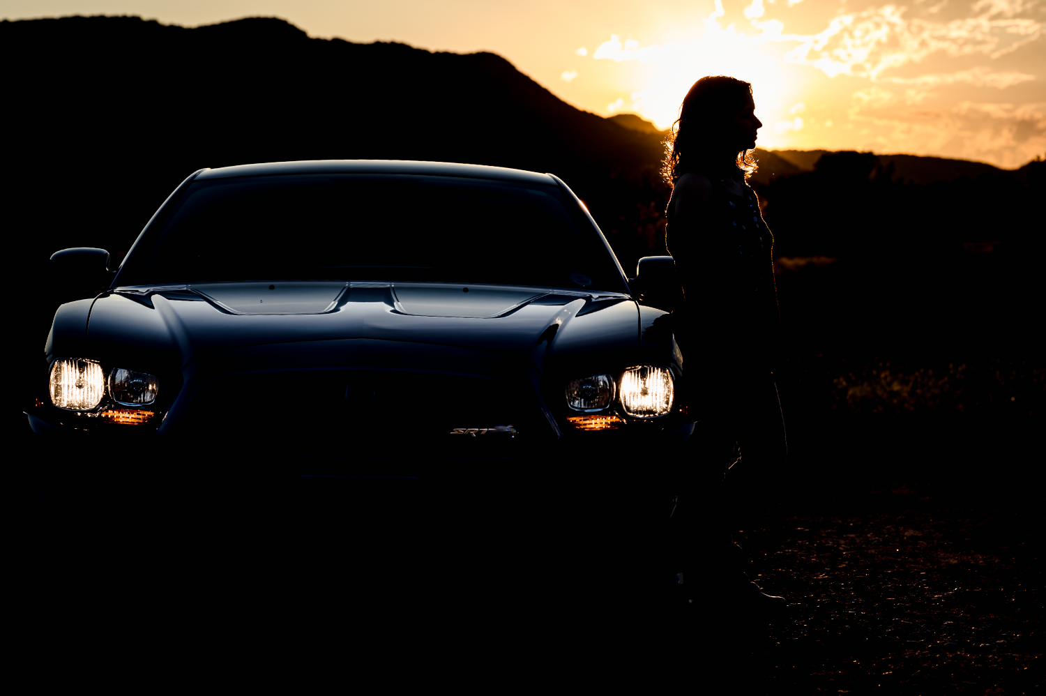 Silhouette of a woman standing next to a dark sports car during sunset, with mountains in the background.