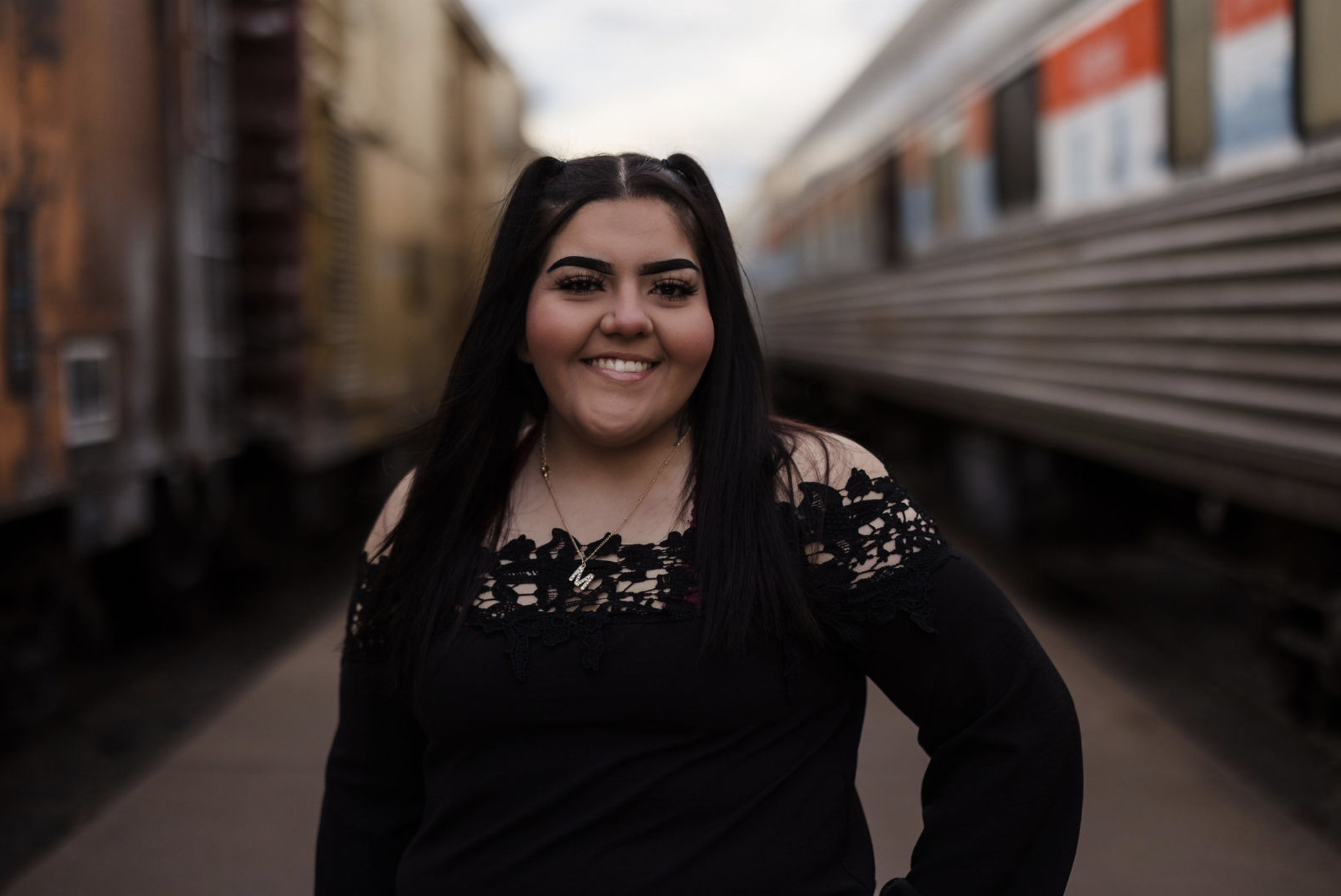 A young woman smiling on a train platform with two trains, one yellow and one silver, in the background.