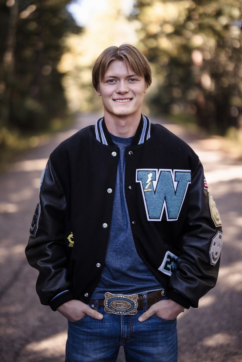 Young man standing outdoors on a dirt path surrounded by trees, smiling, wearing a black varsity jacket with patches and letters, blue t-shirt, blue jeans, and a large decorative belt buckle.
