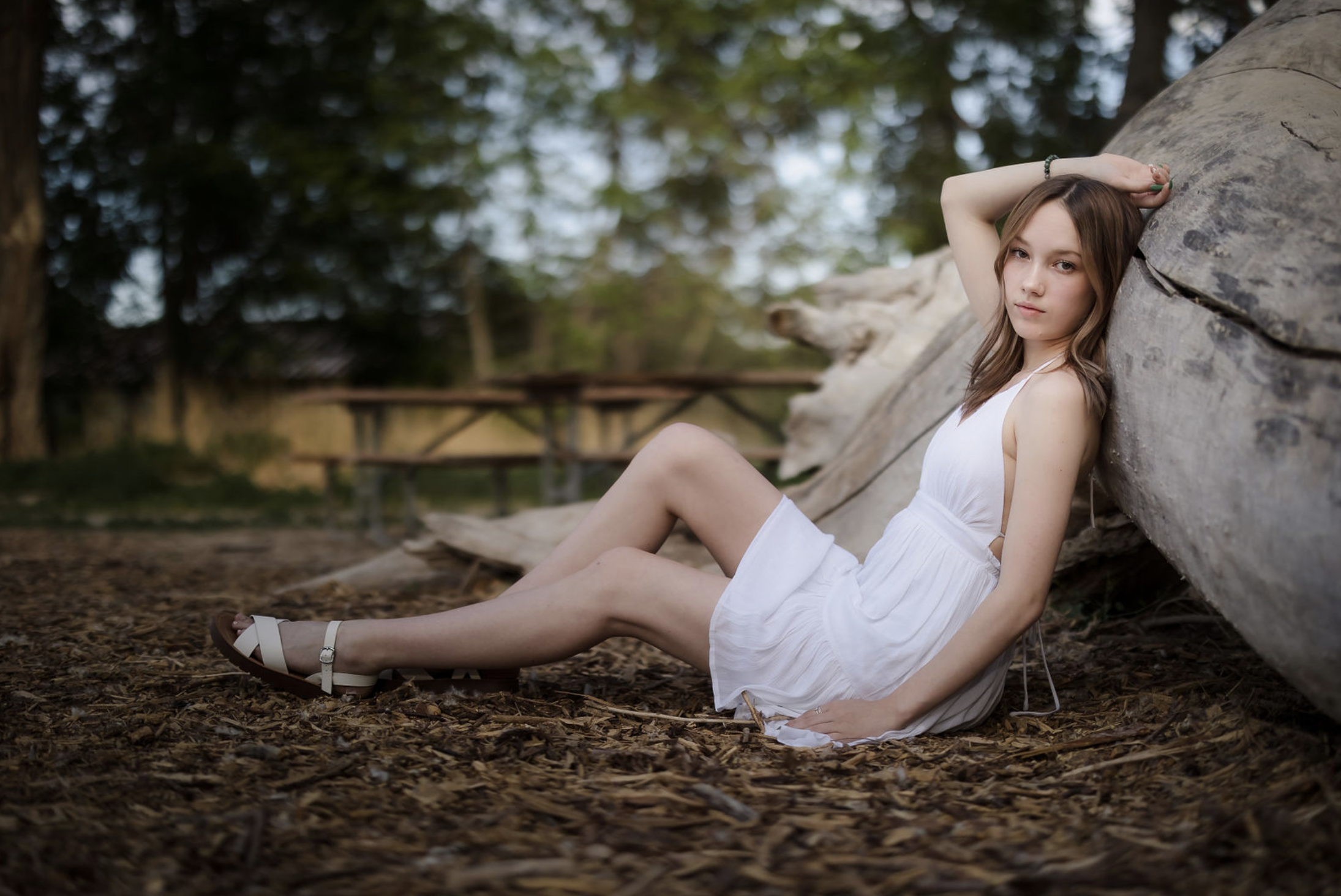 A young woman with curly brown hair, wearing a white dress, sitting on a fallen tree trunk in a lush green forest, smiling at the camera.