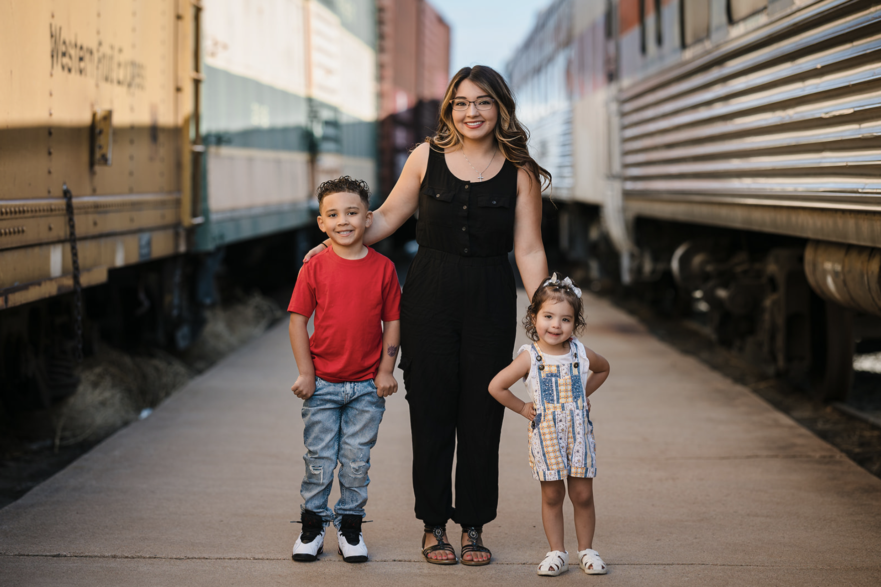 Two children playing near the edge of a lake during sunset, with trees in the background and the sun reflecting on the water.