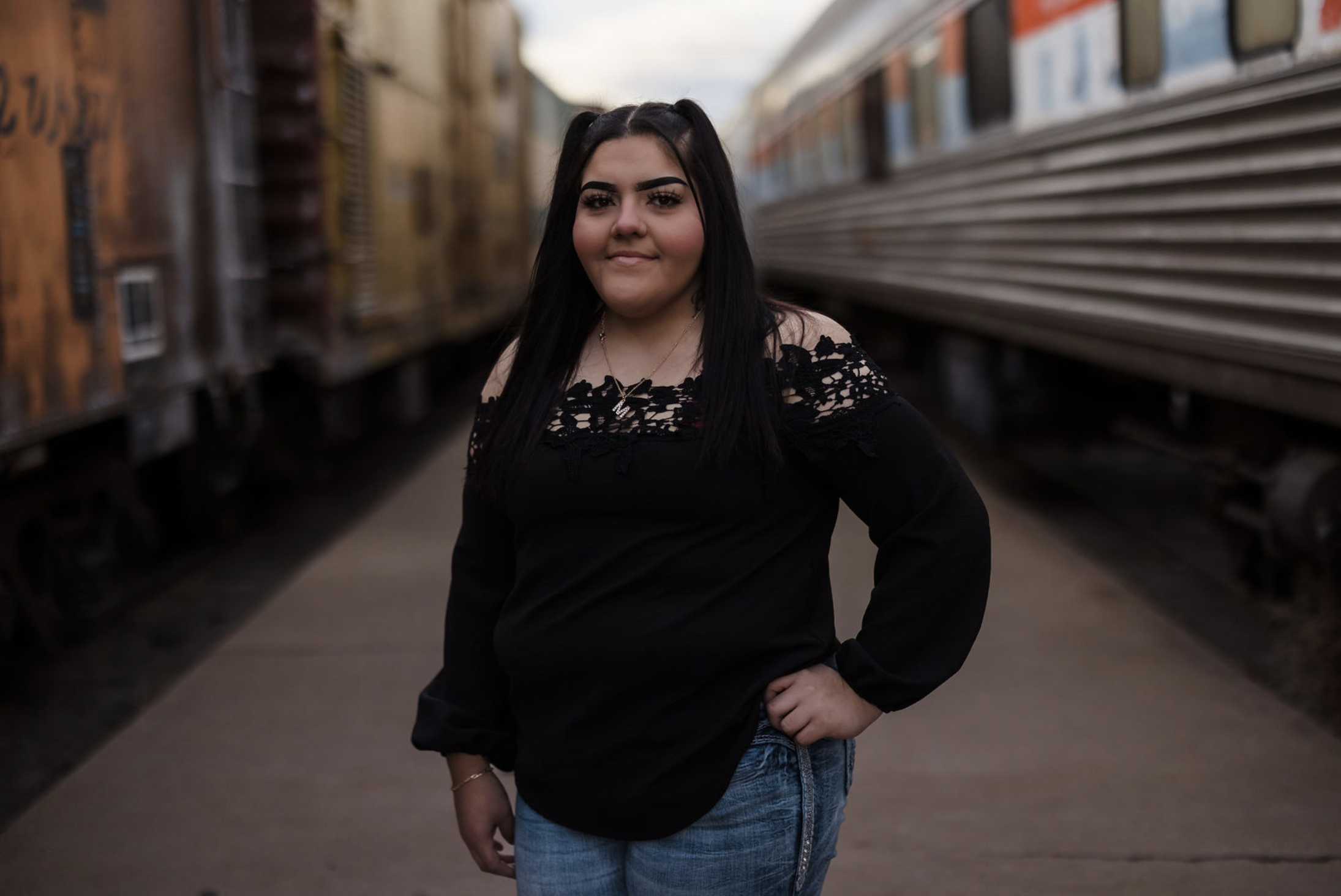 A young girl and boy hugging on a dirt path in a wooded area with autumn leaves, with a man and woman standing in the background, slightly out of focus.