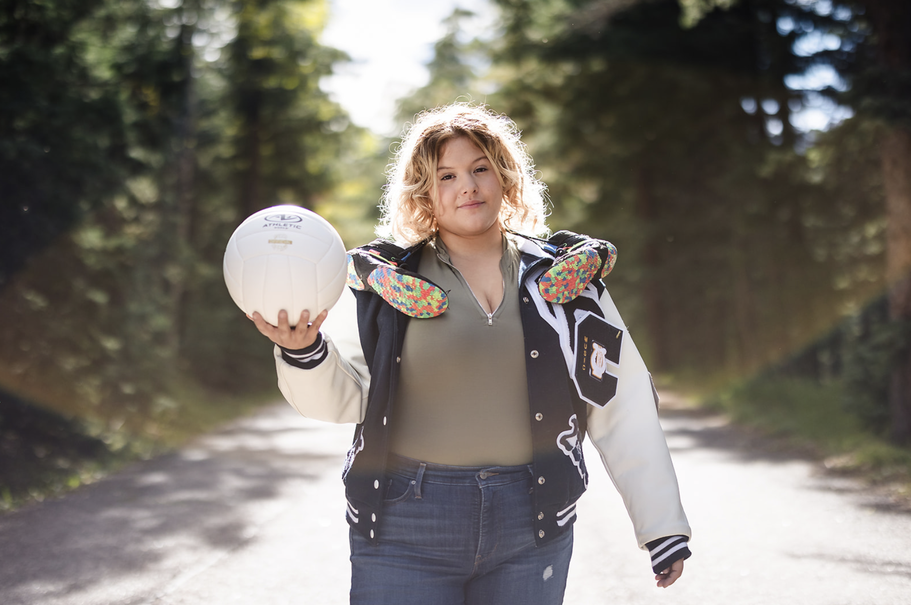 Young woman with curly blonde hair wearing a varsity jacket and carrying a volleyball on her shoulder, standing on a forest trail.