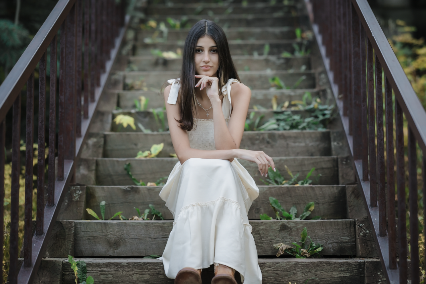 Young woman with long dark hair sitting on wooden outdoor stairs surrounded by greenery, wearing a white dress and brown shoes.