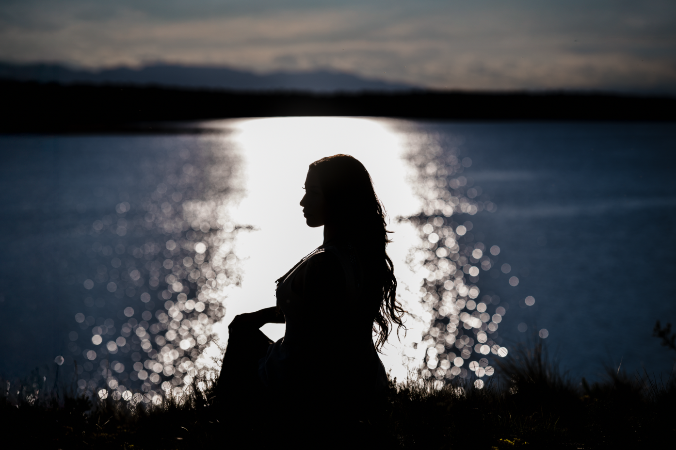 A young girl and boy hugging on a dirt path in a wooded area with autumn leaves, with a man and woman standing in the background, slightly out of focus.
