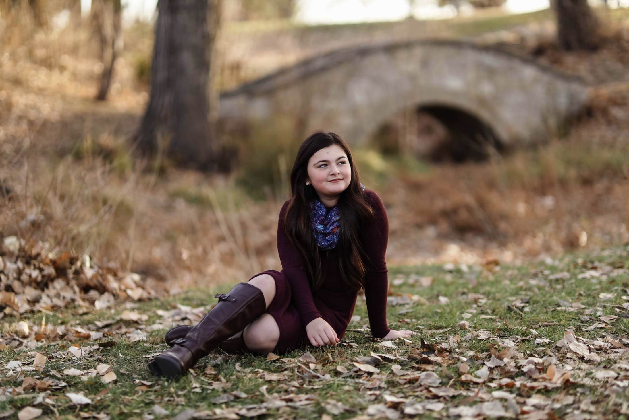 A mature couple stands close to each other by a lake with autumn trees in the background. The woman has gray hair and is wearing a brown sweater with hoop earrings and a necklace. The man has gray hair, glasses, and is wearing a plaid shirt. They are holding hands and looking at the camera.