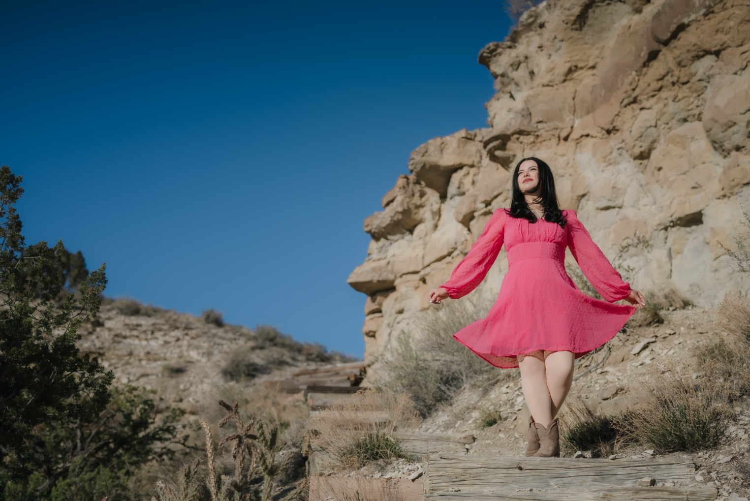 A young girl in a yellow dress with ruffled sleeves and black buttons sitting on a person's shoulder outdoors, smiling and looking at someone out of frame, with trees and greenery in the background.
