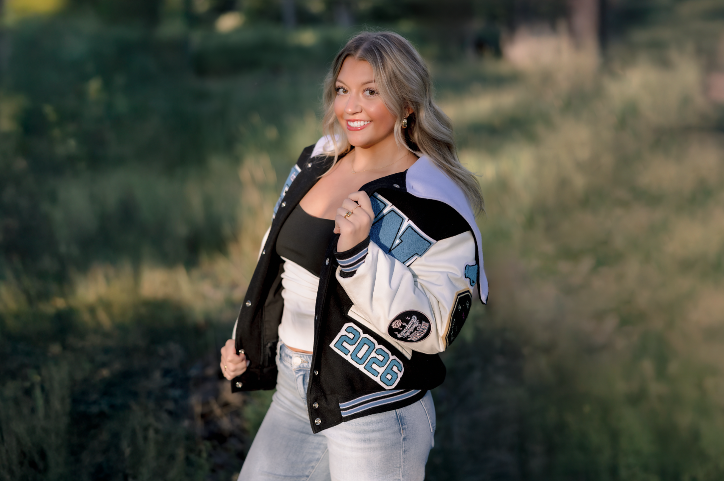 A young woman with blonde wavy hair smiling outdoors near a body of water, wearing a black tank top, a black and white varsity jacket with patches and numbers, and light blue jeans.