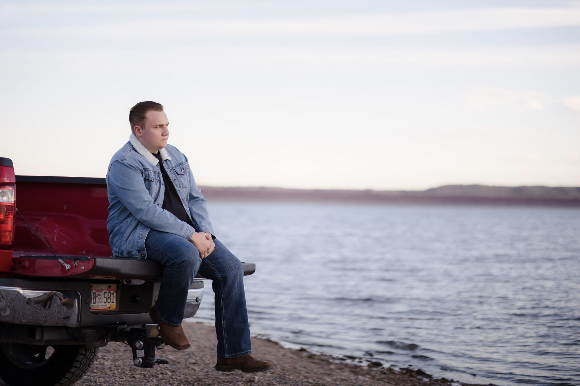 A young man sitting on the tailgate of a pickup truck parked beside a body of water, looking contemplative, with a landscape of water and distant shore in the background.
