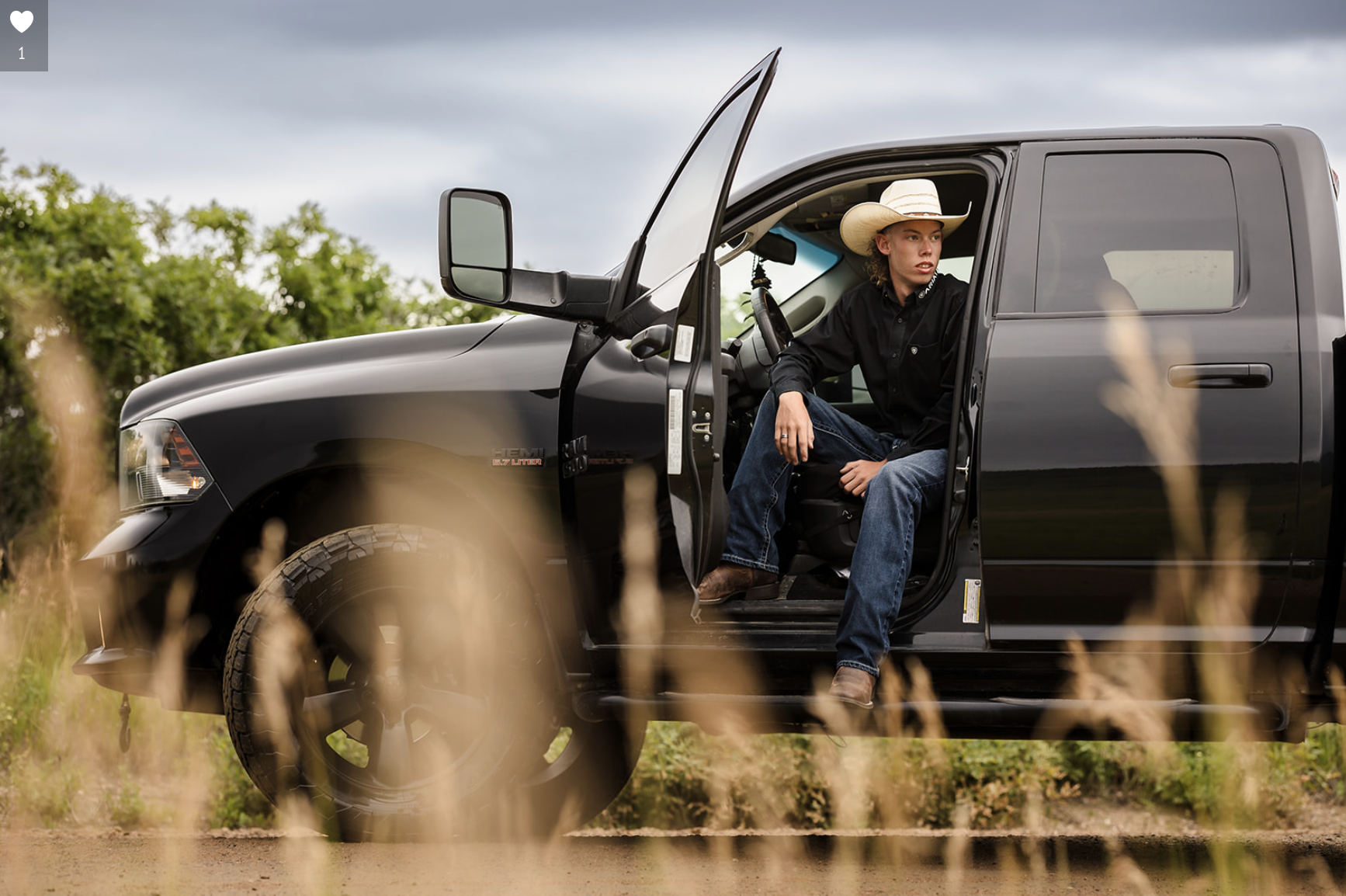 A young man wearing a cowboy hat and black shirt sitting in the driver's seat of a black pickup truck with the door open outdoors, with tall grass in the foreground and green bushes in the background under a cloudy sky.