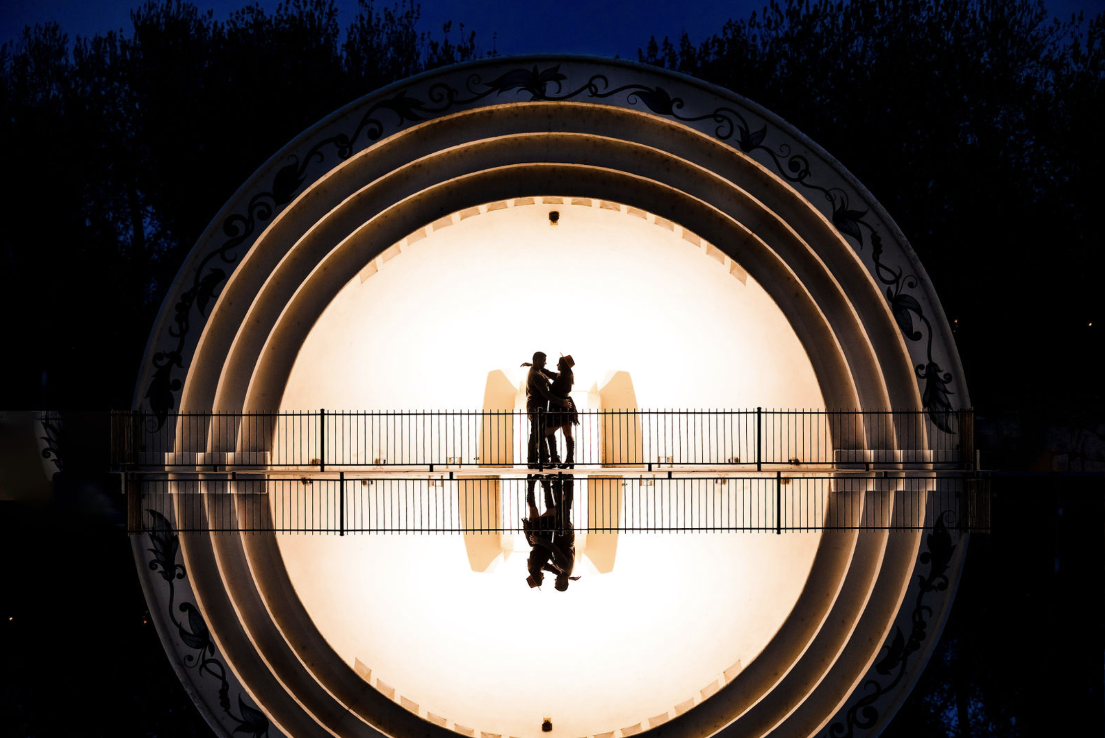 A young girl and boy hugging on a dirt path in a wooded area with autumn leaves, with a man and woman standing in the background, slightly out of focus.