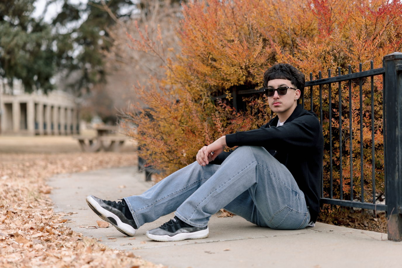 A young woman with dark brown wavy hair and blue eyes smiling outdoors in a park during daytime, wearing a white sweater and layered gold necklaces.