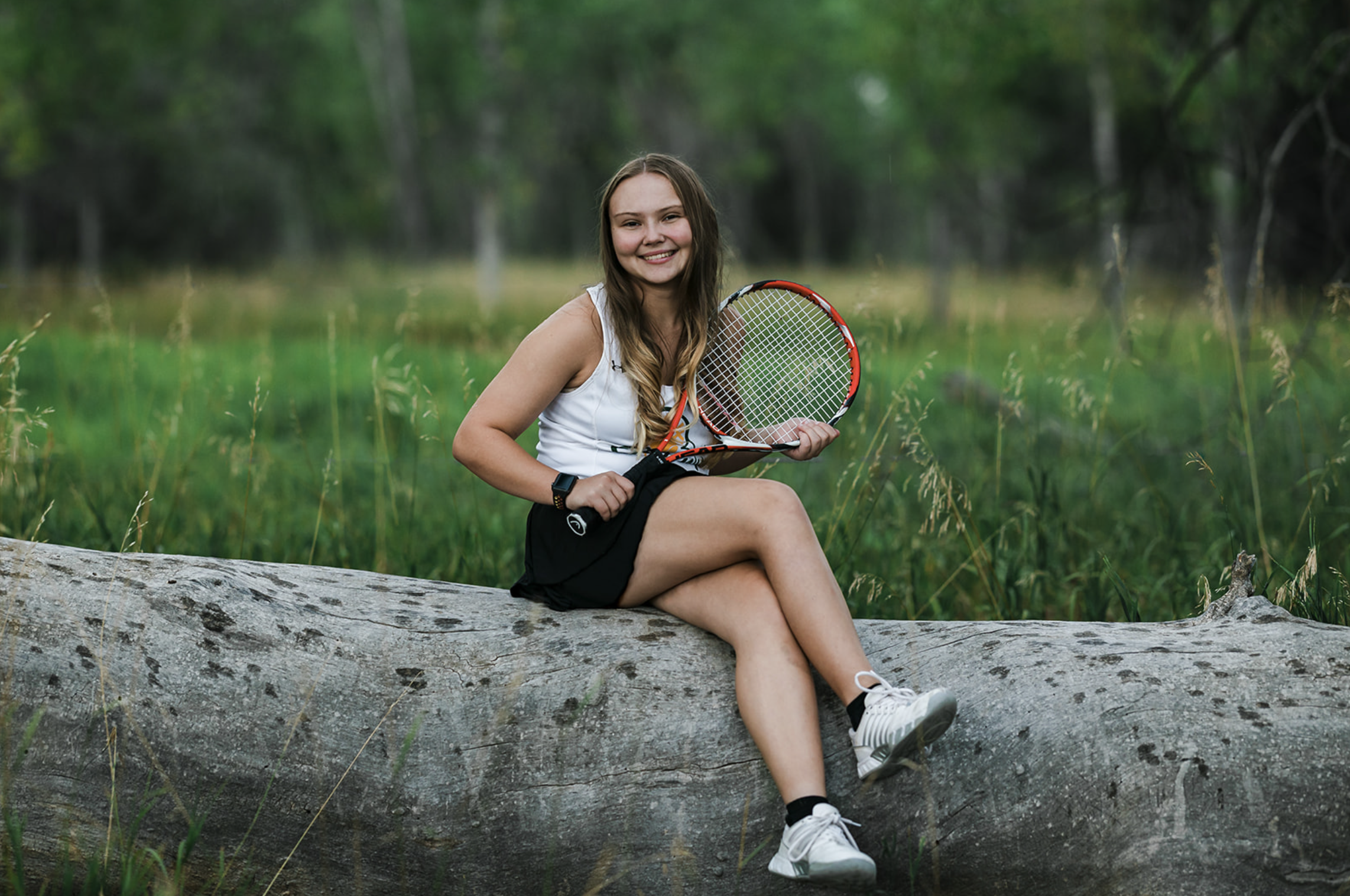 A young woman sitting on a fallen tree in a grassy outdoor area, holding tennis rackets, smiling at the camera.