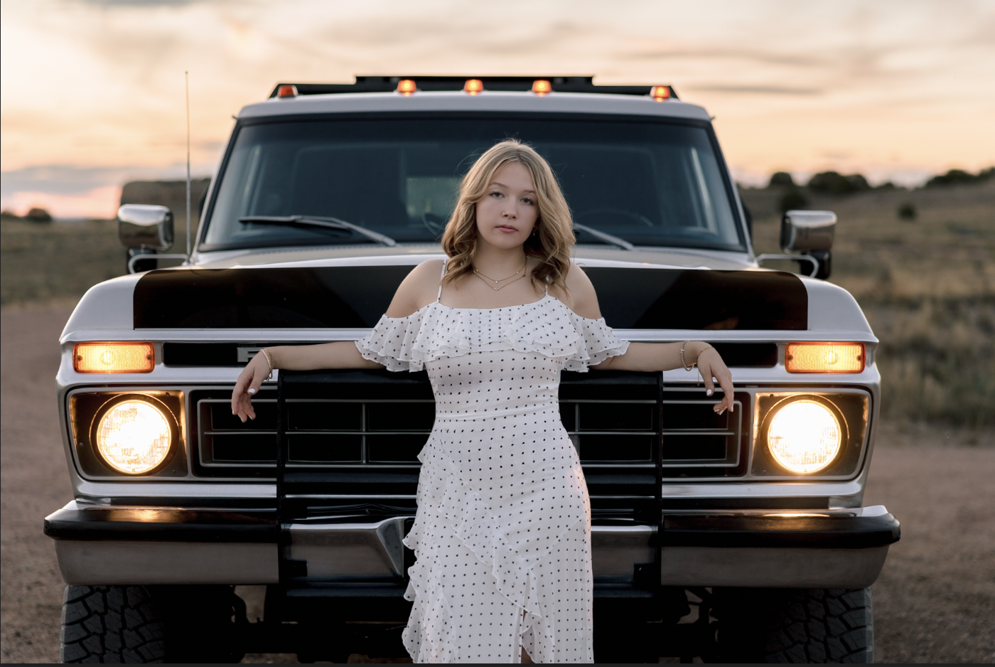 A young woman with wavy blonde hair, wearing a white polka dot dress, stands in front of a black and white SUV with sunlight behind her at sunset.