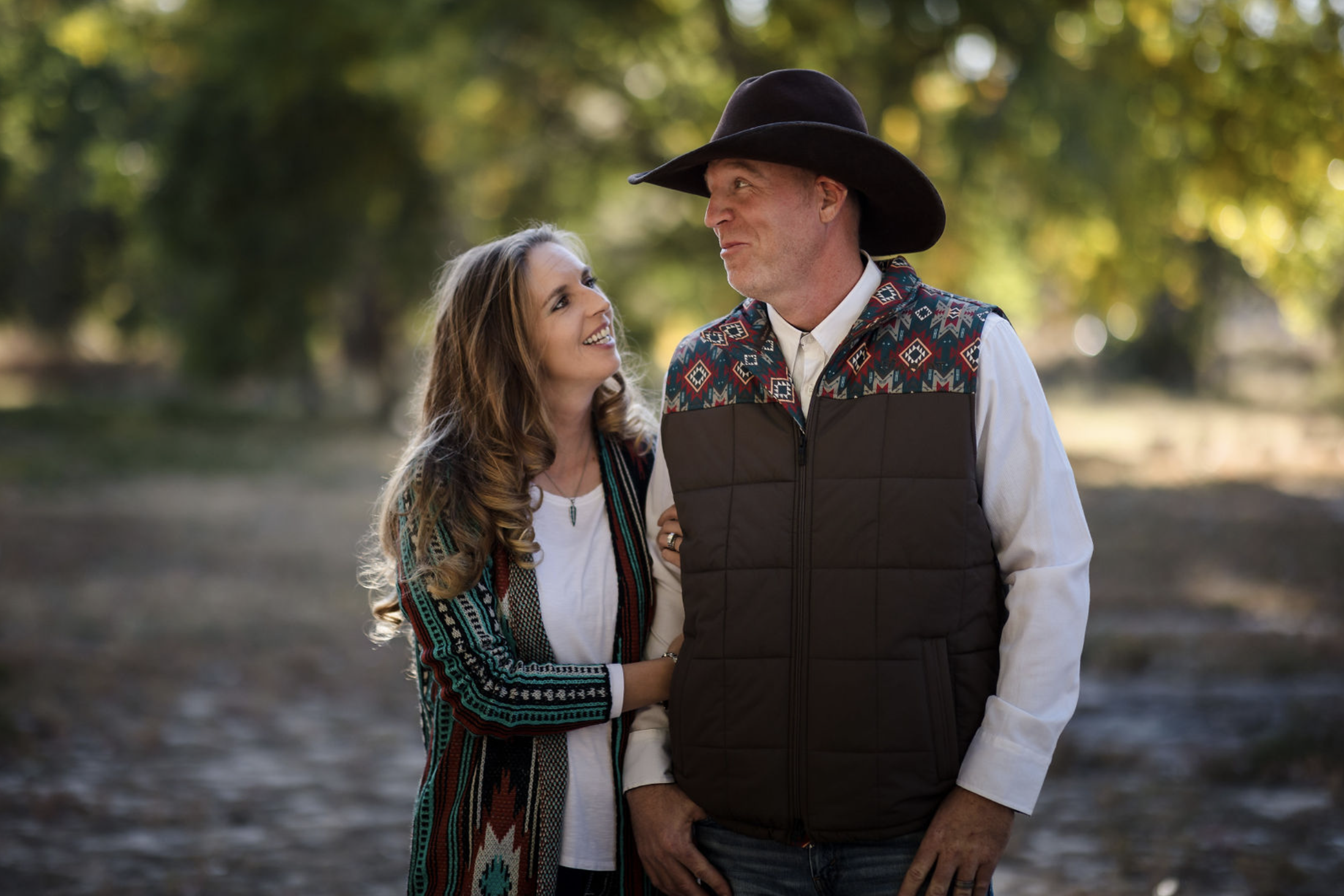 A young woman with dark brown wavy hair and blue eyes smiling outdoors in a park during daytime, wearing a white sweater and layered gold necklaces.