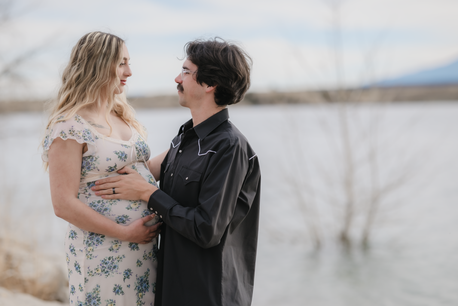 A young girl in a yellow dress with ruffled sleeves and black buttons sitting on a person's shoulder outdoors, smiling and looking at someone out of frame, with trees and greenery in the background.