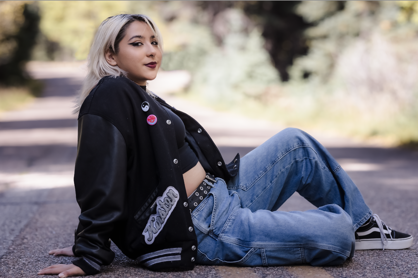 A young woman with platinum blonde hair, wearing a black jacket with patches, a black crop top, blue jeans, and black and white sneakers, sitting on an empty road with a background of trees and autumn foliage.