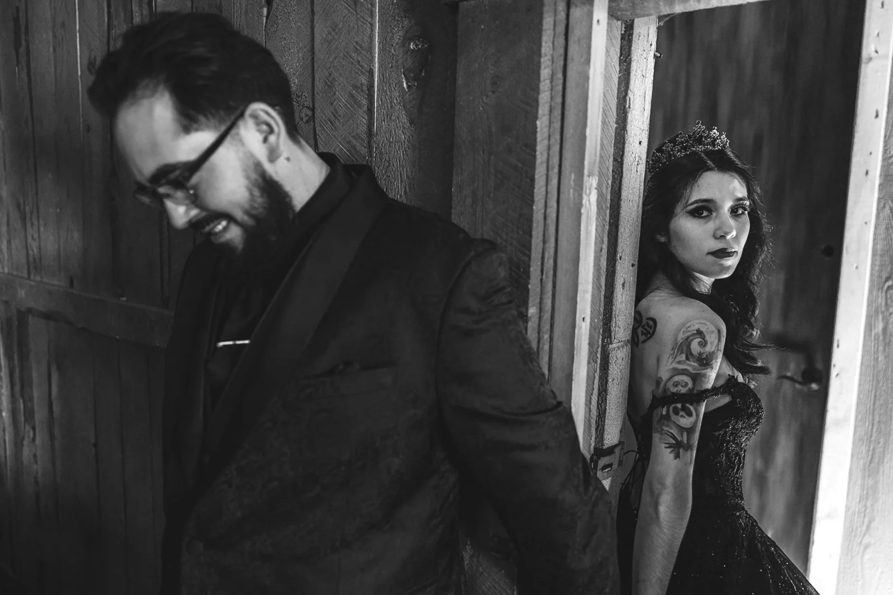 A man and a woman are standing back to back. The man is smiling and wearing glasses, a black jacket, and a shirt. The woman has dark hair, tattoos on her arm, dark makeup, and is wearing a black dress and a tiara, looking at the camera with a serious expression.