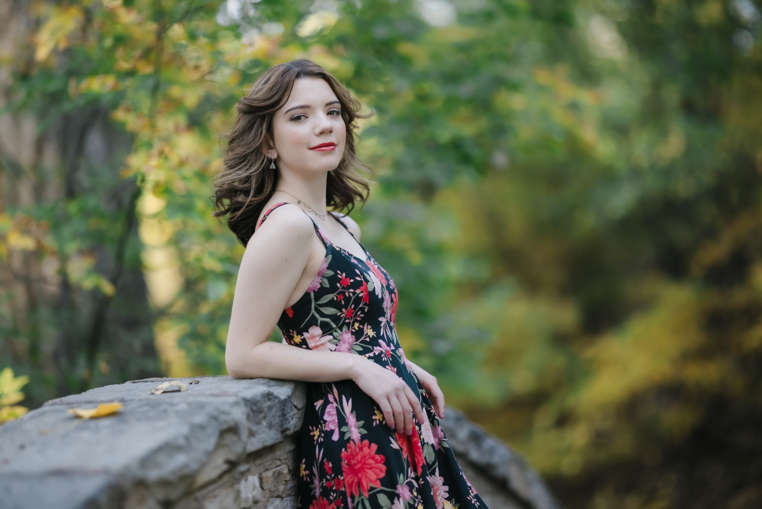 A young woman with dark brown wavy hair and blue eyes smiling outdoors in a park during daytime, wearing a white sweater and layered gold necklaces.