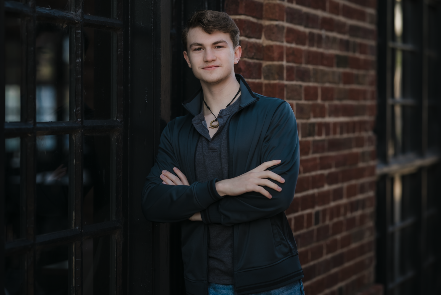 Young man with short brown hair wearing a dark blue jacket and gray shirt, standing with arms crossed against a brick wall and black metal window frame, outdoors.
