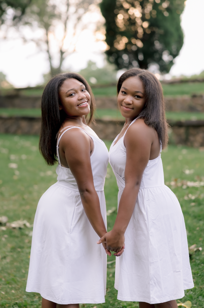 Two women standing on grass, holding hands, smiling, wearing white dresses in an outdoor park with trees in the background.