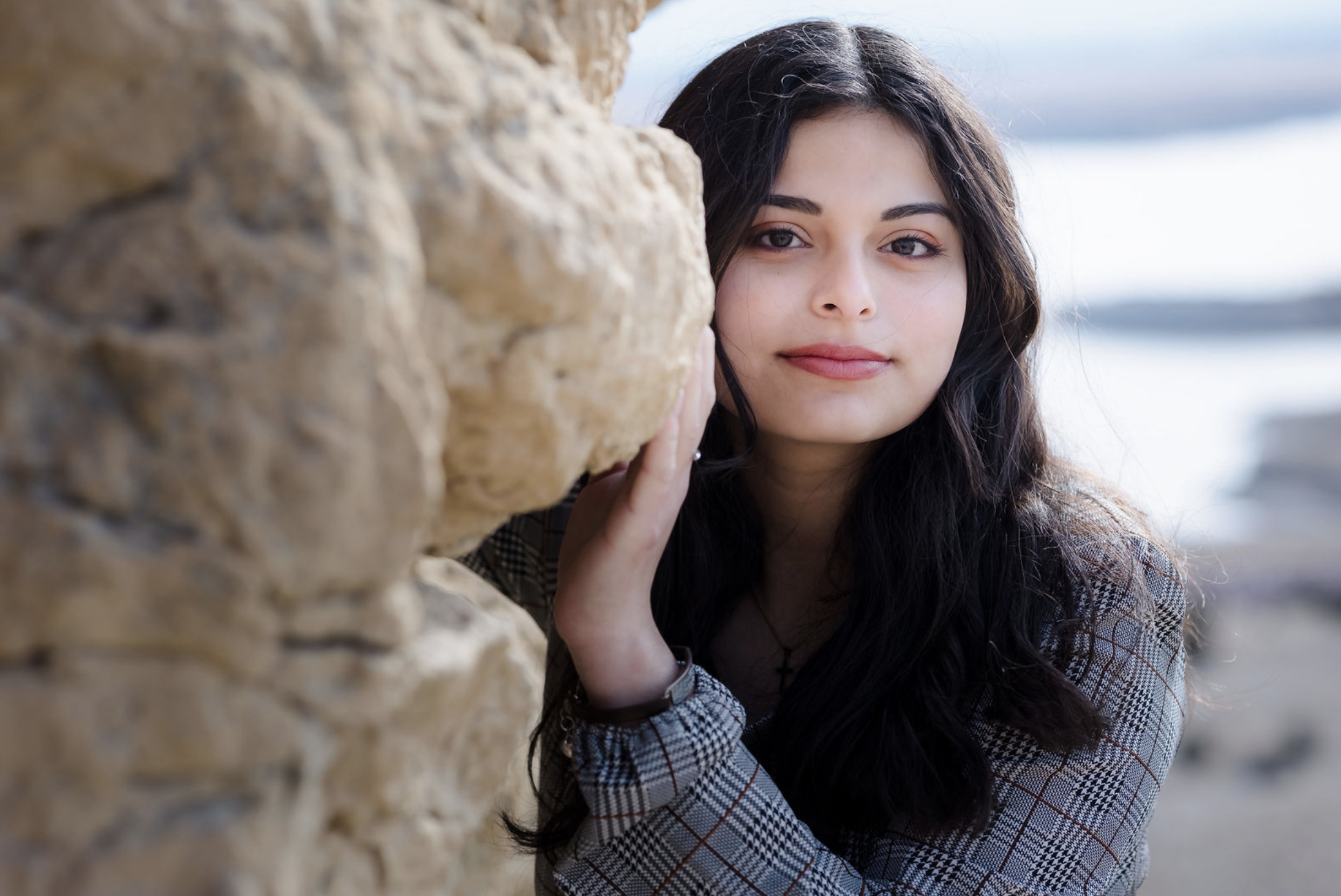 A young man with tousled blond hair and a serious expression, wearing a light gray T-shirt with buttons, standing outdoors in front of a calm body of water and a blurred green landscape.