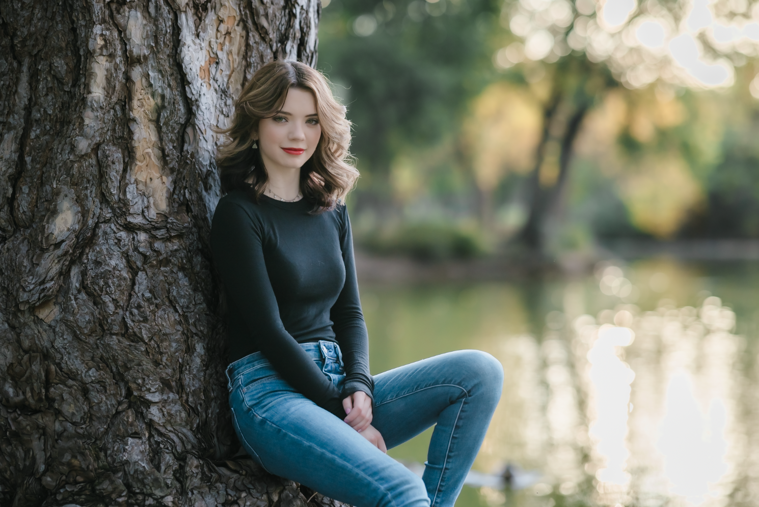 A young woman with wavy brown hair, wearing a black long-sleeve shirt and blue jeans, sitting by a large tree near a body of water during late afternoon or early evening, with a blurred background of green trees and water reflects sunlight.