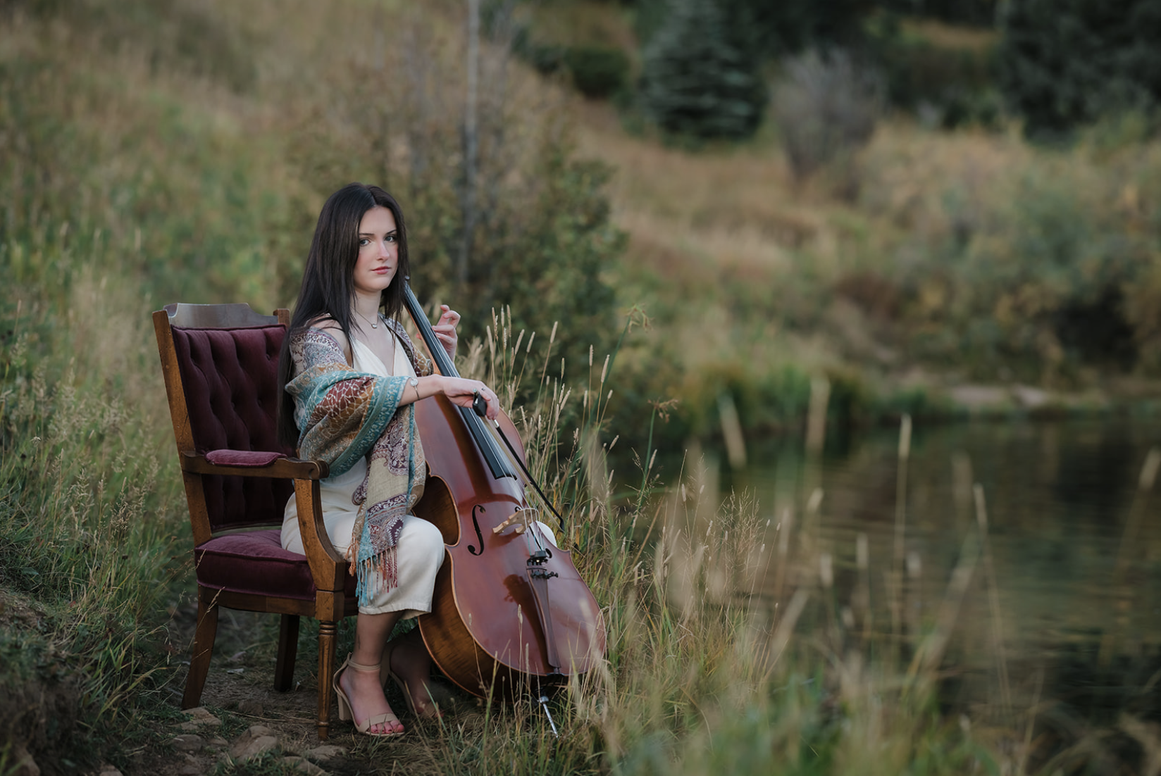 A woman with long dark hair playing a cello outdoors near a body of water, seated on a vintage chair with a velvet cushion, surrounded by tall grass and trees.