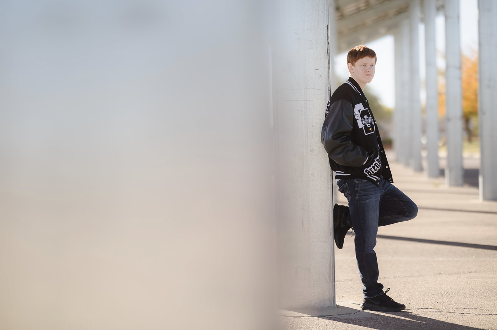 A young man with red hair, wearing a black and gray varsity jacket and jeans, leaning against a white concrete wall under a bridge.