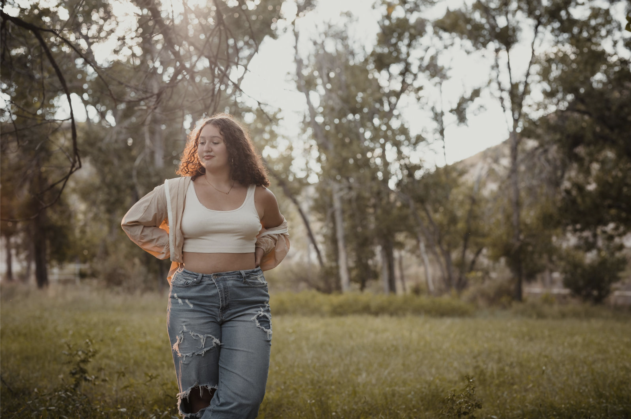 A young woman with curly hair wearing a beige jacket, white crop top, and ripped jeans walking outdoors in a grassy area with trees in the background, illuminated by warm sunlight.