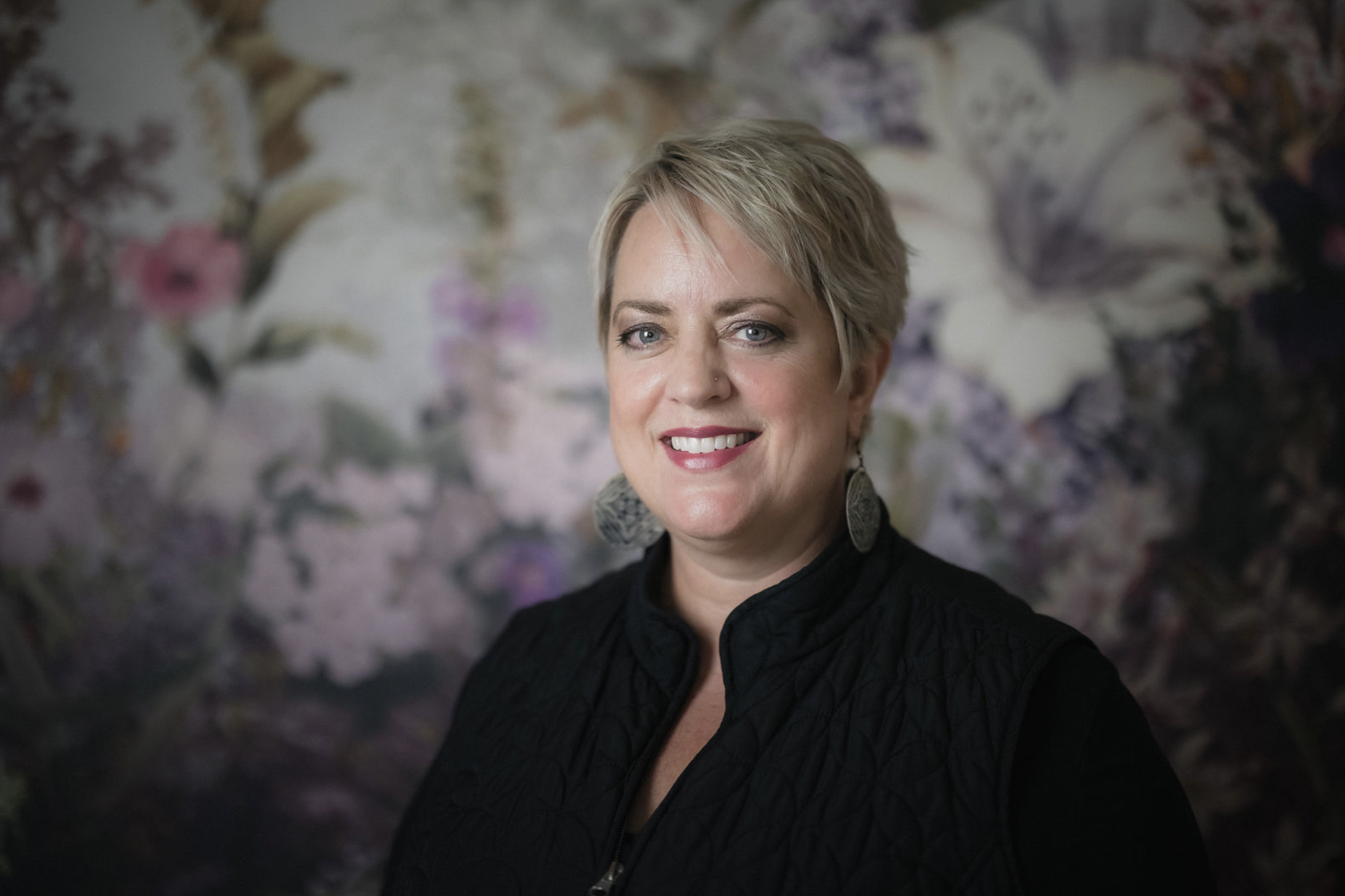 A young woman with wavy blonde hair, blue eyes, and light skin, wearing a black top and a gold necklace, sitting outdoors at a table with her arms crossed, looking at the camera with a neutral expression.