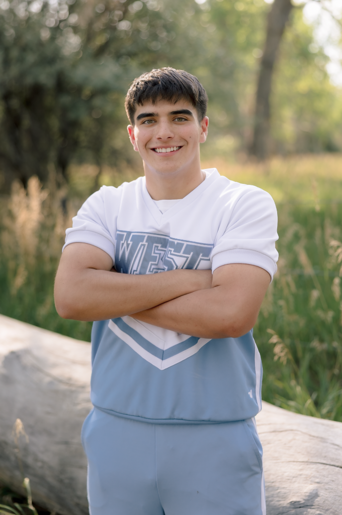 A young man smiling outdoors with arms crossed, standing near a fallen log in a grassy field with trees in the background.