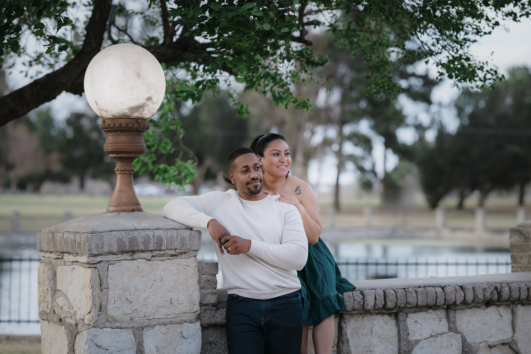 Young man sitting outdoors on grass near a tree, wearing a colorful floral shirt and blue jeans, with a thoughtful expression.