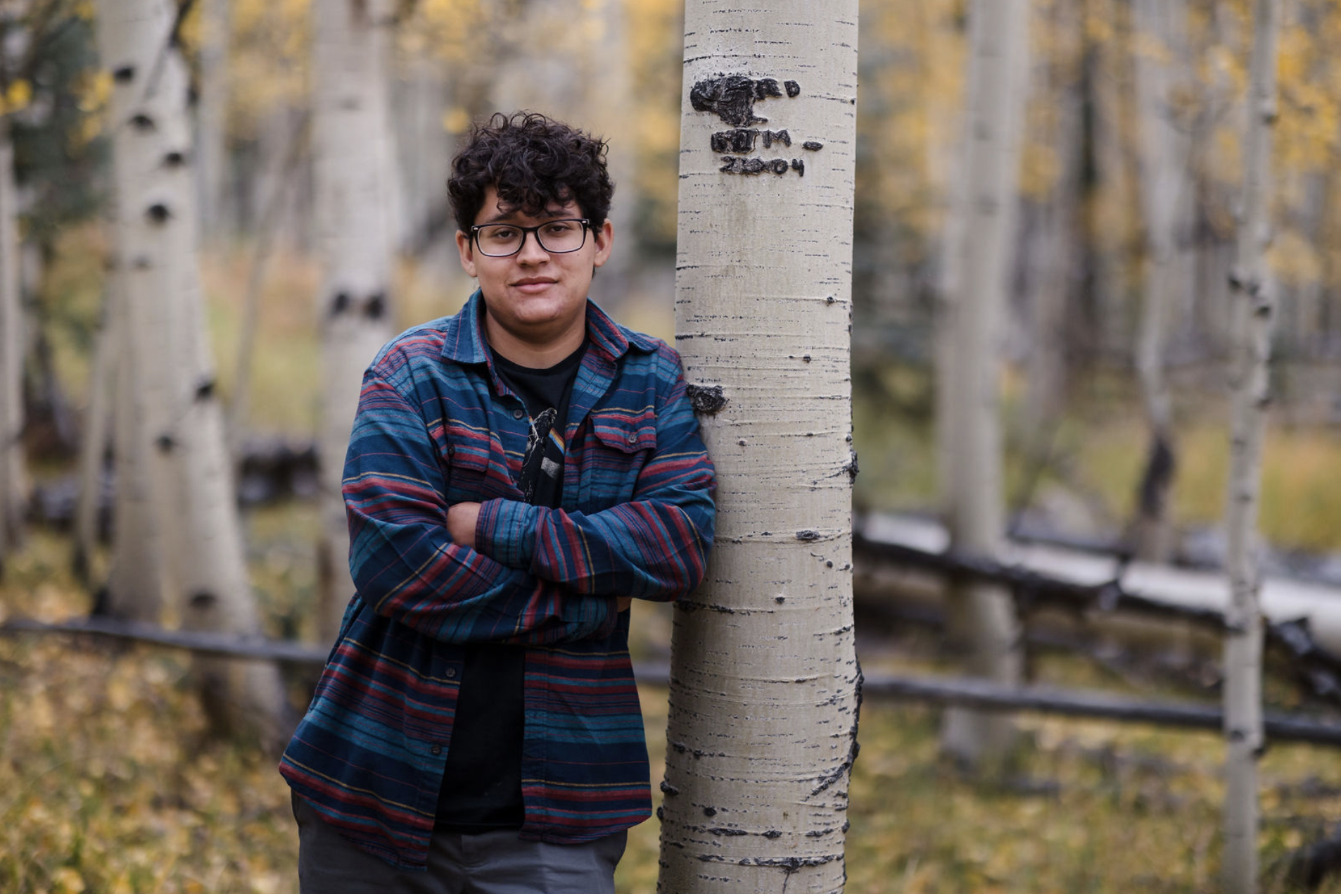 A young man with glasses and curly hair stands with crossed arms beside a birch tree in a forest during autumn.