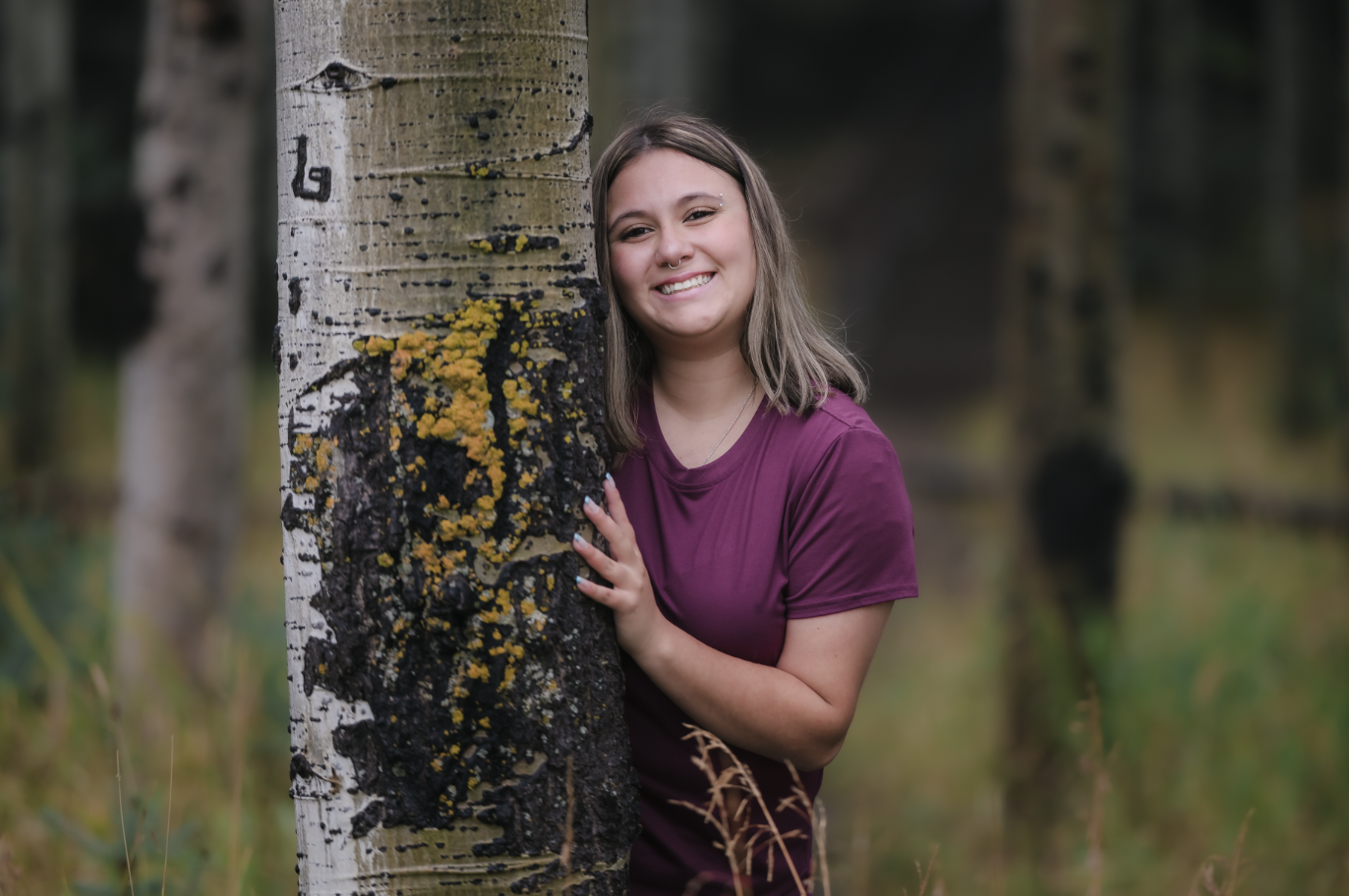 A young woman with shoulder-length brown hair, smiling, standing behind a tall birch tree with yellow and black mold on the trunk, in a forest setting.