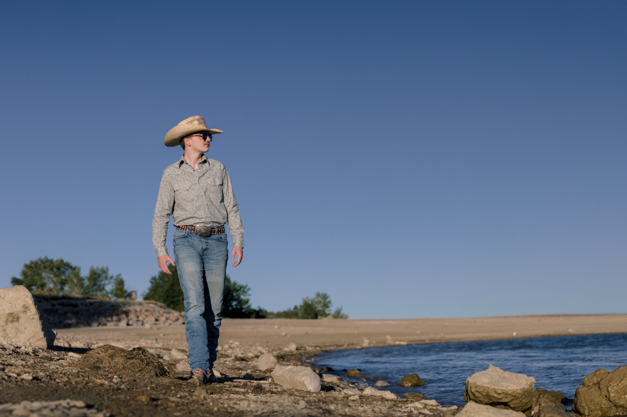 Woman in cowboy hat, sunglasses, and western attire walking on a rocky beach near water under clear blue sky.