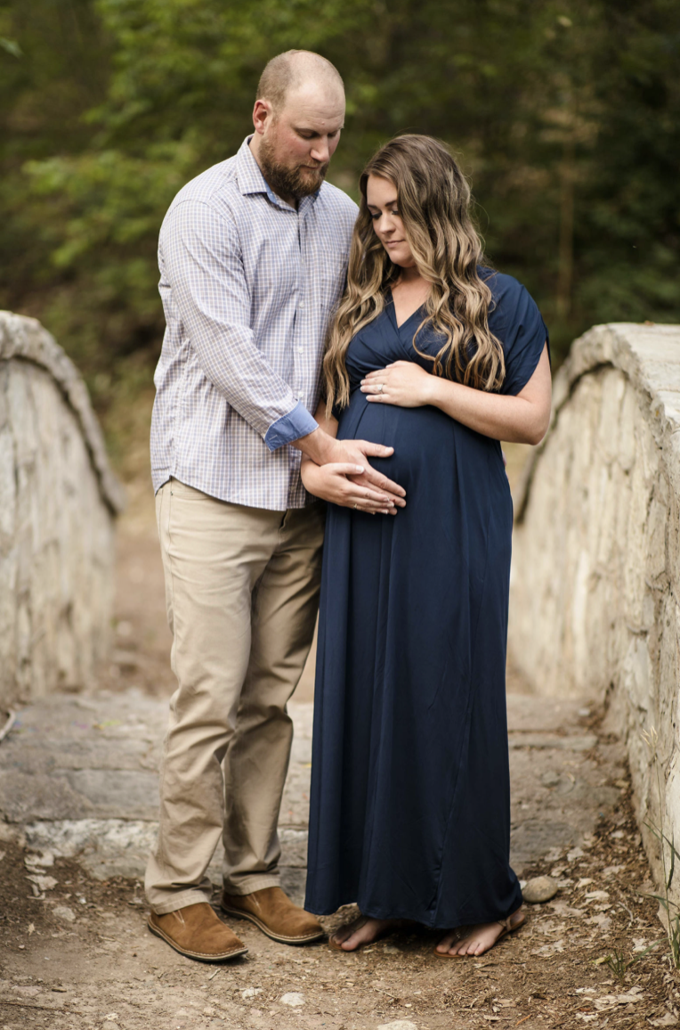 A young girl in a yellow dress with ruffled sleeves and black buttons sitting on a person's shoulder outdoors, smiling and looking at someone out of frame, with trees and greenery in the background.