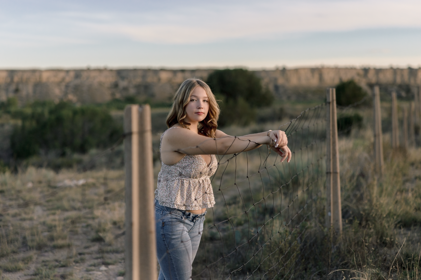 A young woman with shoulder-length wavy hair, wearing a floral sleeveless top and jeans, standing behind a wire fence in a rural landscape during sunset, with a canyon or cliff in the background.