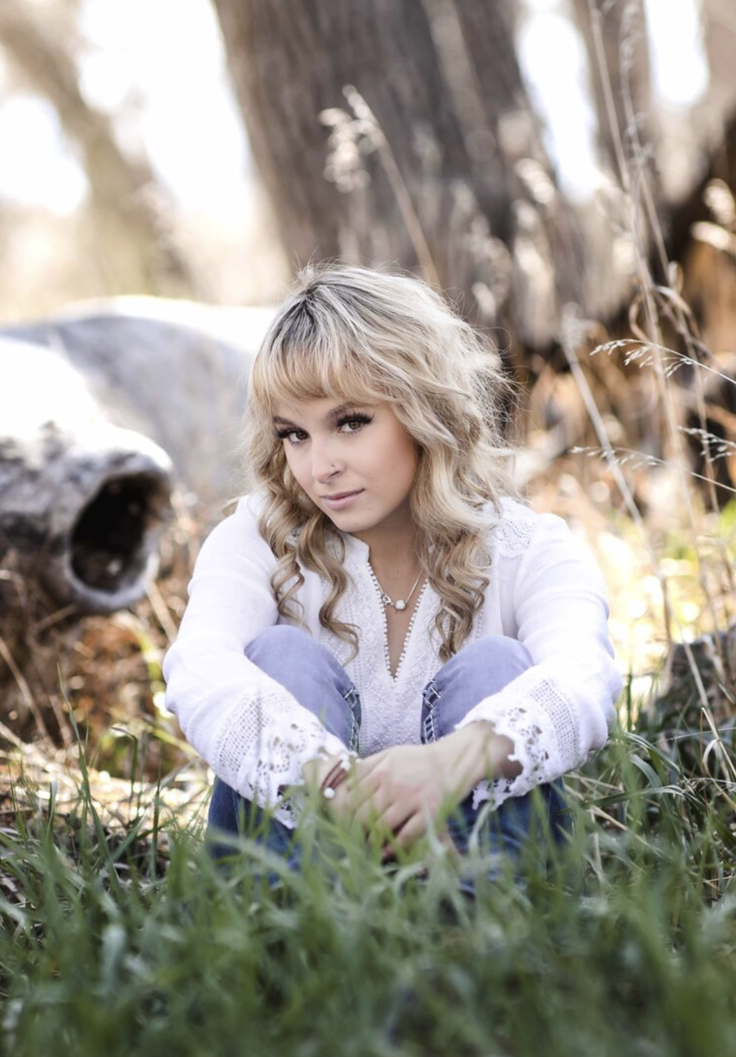 A young man with tousled blond hair and a serious expression, wearing a light gray T-shirt with buttons, standing outdoors in front of a calm body of water and a blurred green landscape.