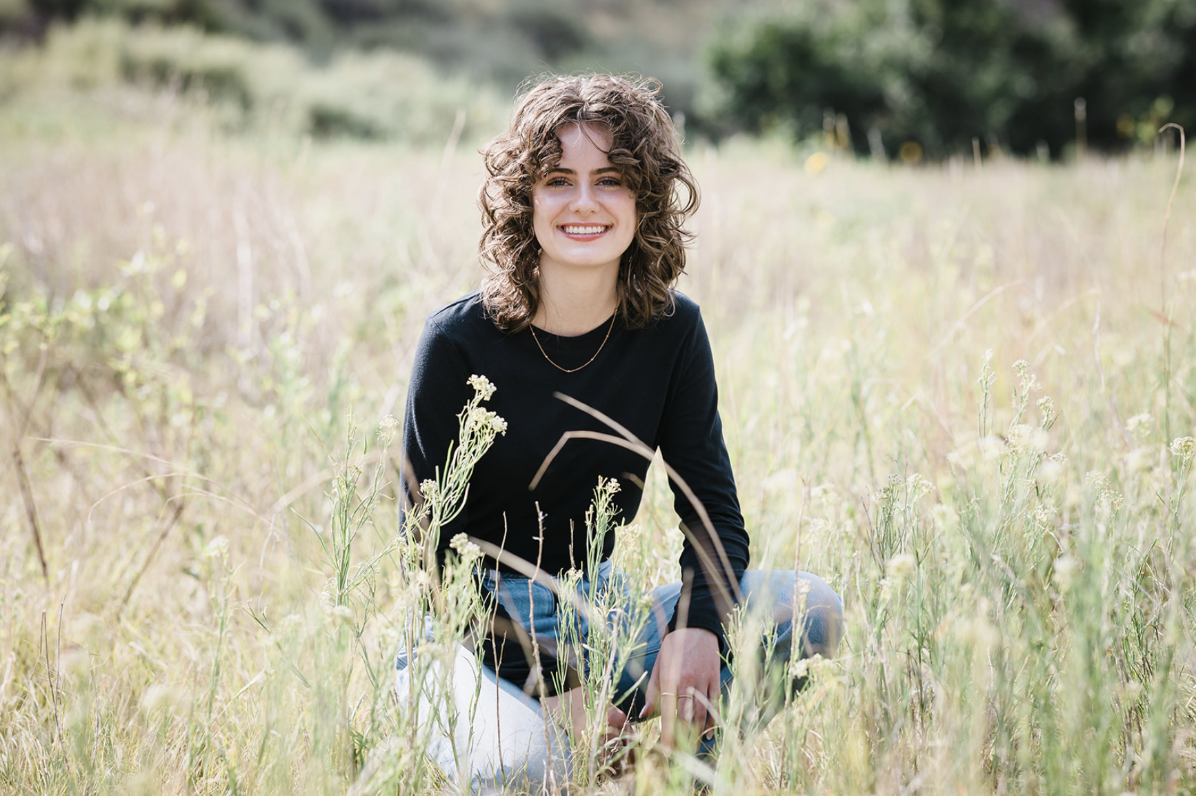 A young woman with curly hair and a black long-sleeve shirt sitting in a grassy field with wildflowers, smiling at the camera.