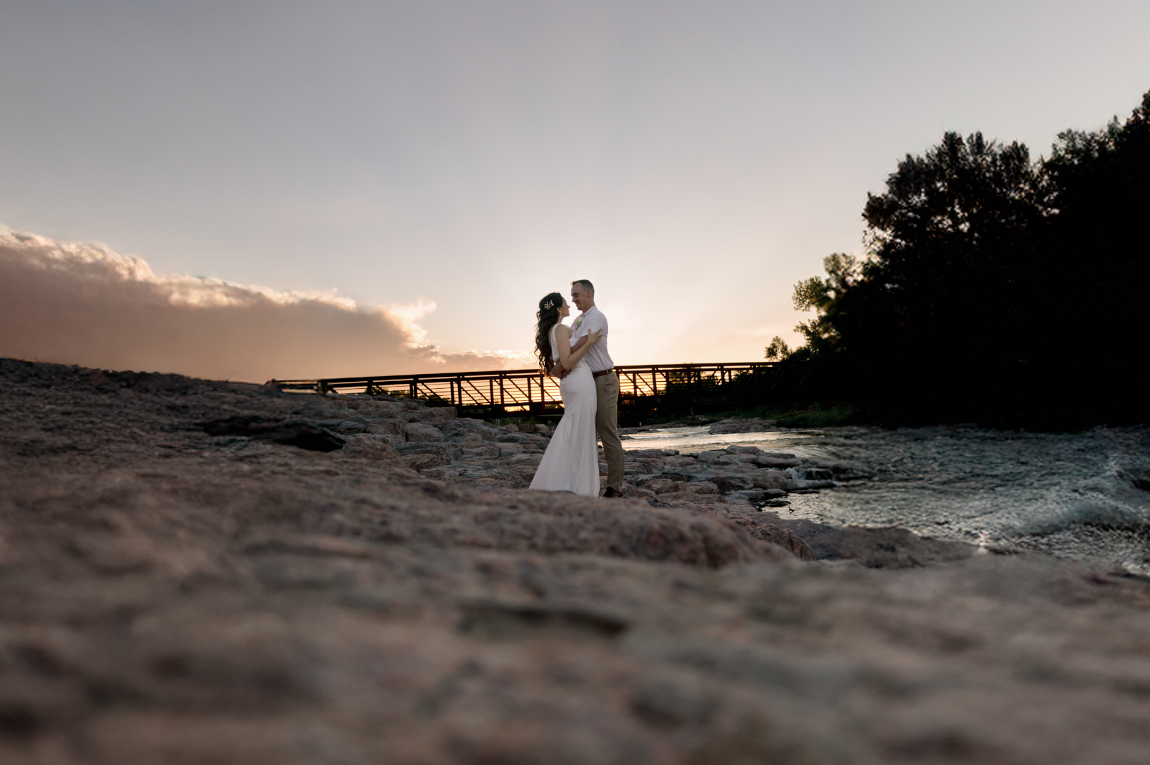 A young woman with dark brown wavy hair and blue eyes smiling outdoors in a park during daytime, wearing a white sweater and layered gold necklaces.