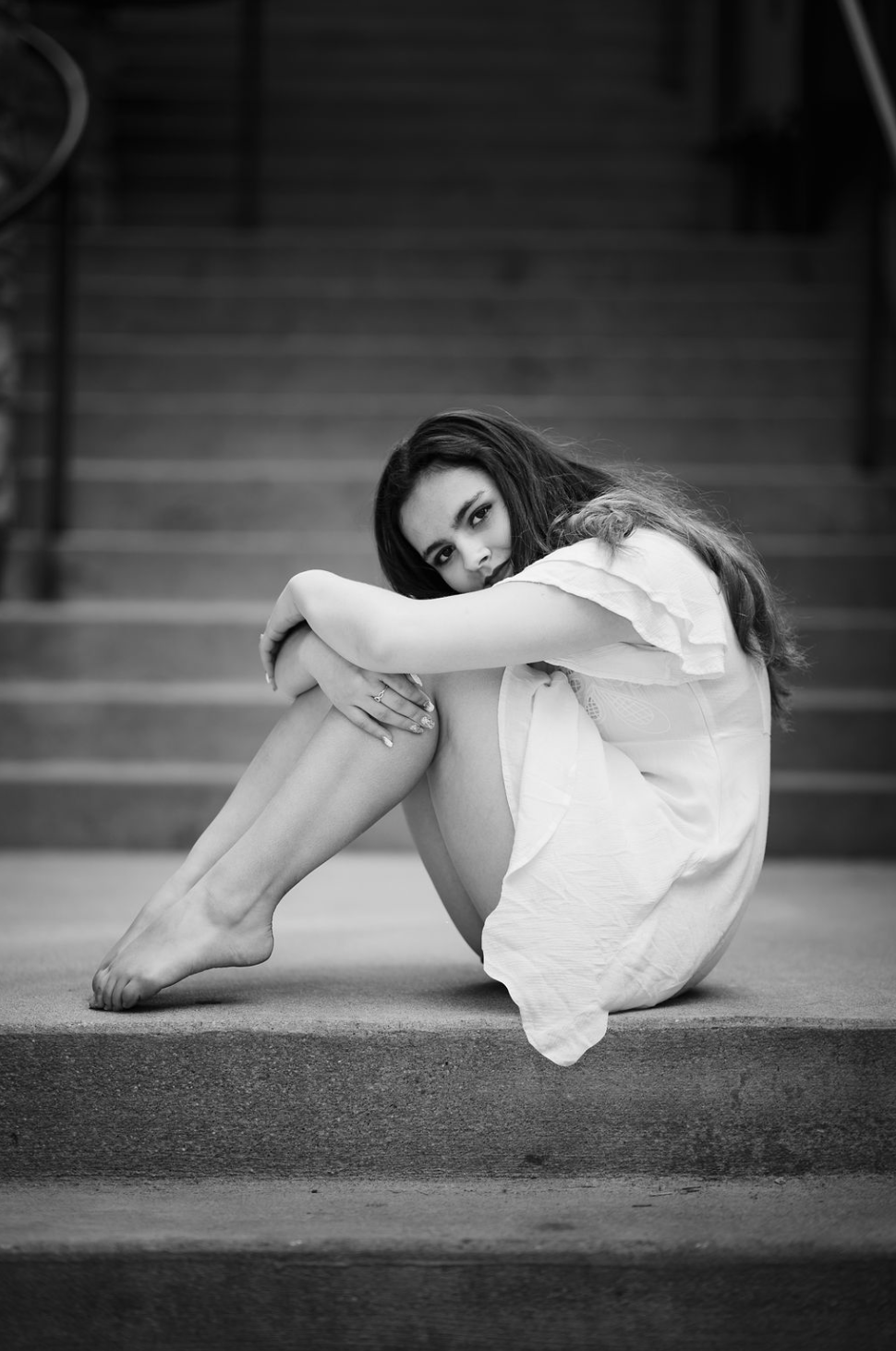 A young man with tousled blond hair and a serious expression, wearing a light gray T-shirt with buttons, standing outdoors in front of a calm body of water and a blurred green landscape.
