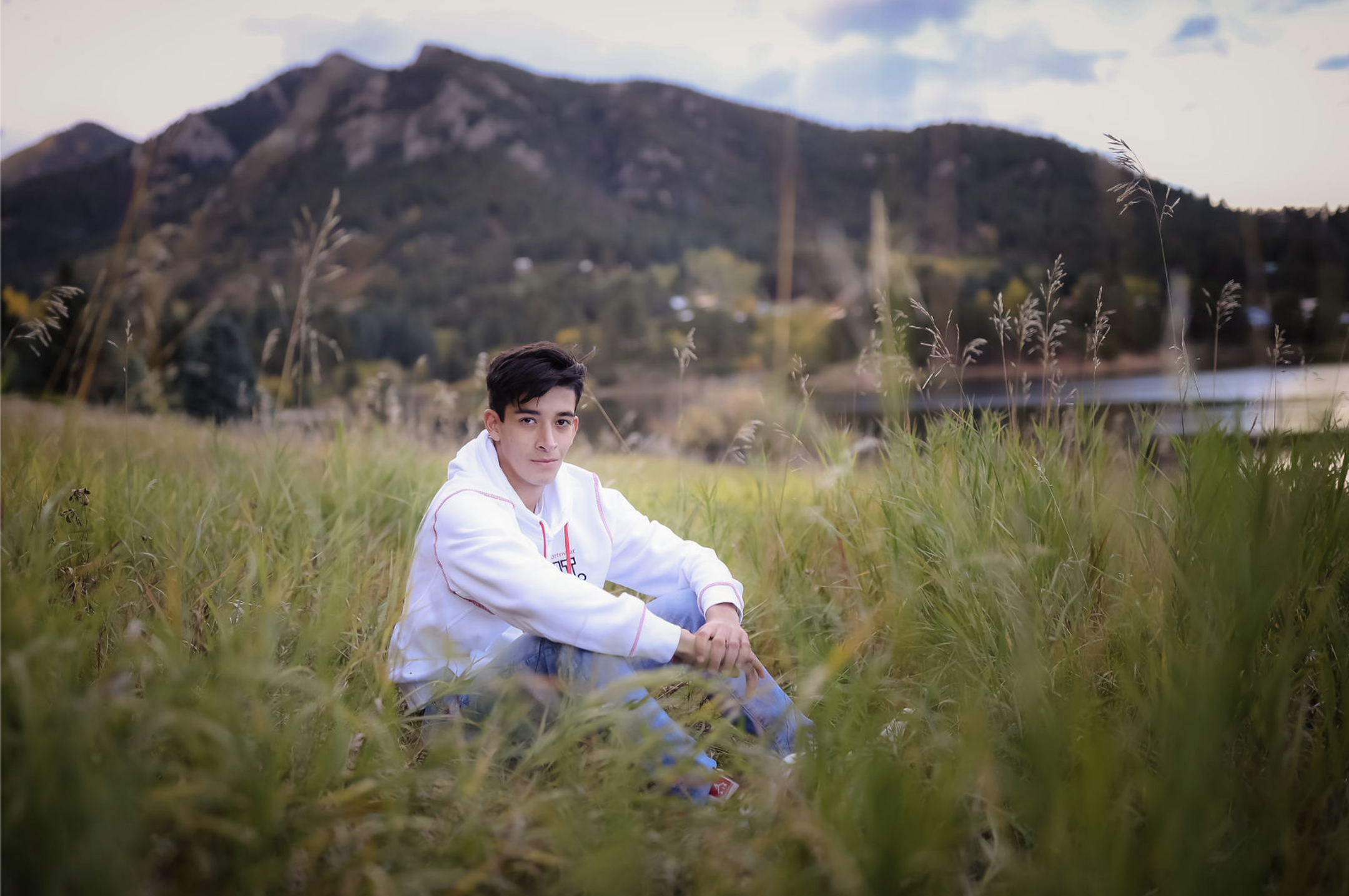 A young man in a white hoodie sitting in tall grass near a body of water with mountains and a cloudy sky in the background.